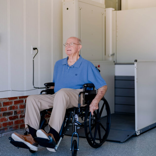A smiling older man in a wheelchair on a Bruno vertical platform lift at a home entrance